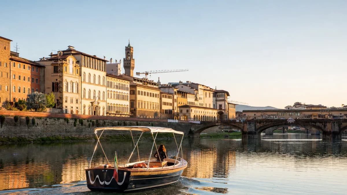 Arno Boat Tour: a small sightseeing boat on the water in Florence, Italy, with the city's real waterfront behind it
